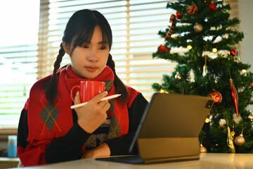 Pretty young woman wearing warm sweater drinking coffee and reading email on digital tablet
