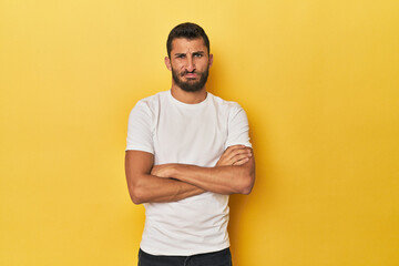 Young Hispanic man on yellow background frowning face in displeasure, keeps arms folded.