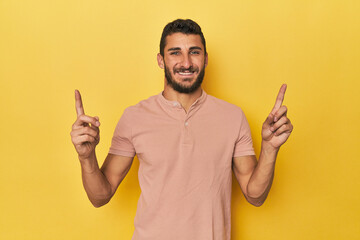 Young Hispanic man on yellow background indicates with both fore fingers up showing a blank space.