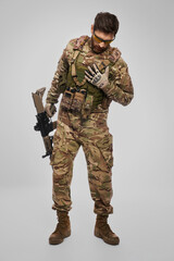 Weak soldier in camouflage outfit covering wound with hand indoors. Front view of wounded bearded military man checking chest, is there an injury, on gray background. Injury, rescue concept.