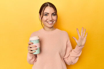 Young caucasian woman holding a takeaway coffee cup smiling cheerful showing number five with fingers.