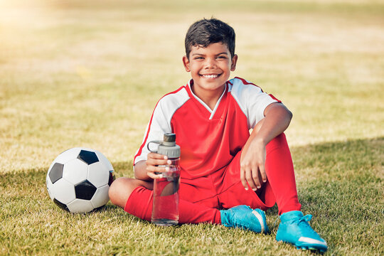Soccer, children and water with a ball and boy child sitting on a grass pitch or field after training. Football, fitness and hydration with a young male kid at a sports venue, stadium or arena
