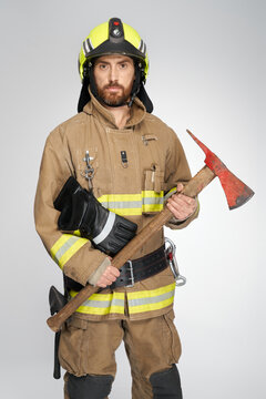 Bearded caucasian firefighter in full gear holding red axe for fire fighting in studio. Front view of handsome fireman with hatchet looking at camera, on gray background. Concept of work, fire tools.