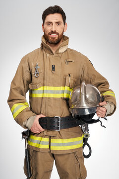Handsome caucasian firefighter smiling to camera indoors. Portrait of happy, strong male fireman in protective uniform holding helmet, on white studio background. Workplace, safety concept.