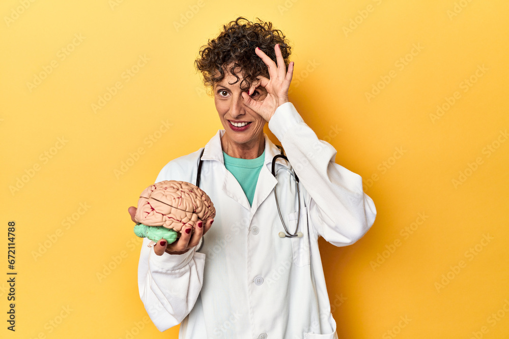 Wall mural Doctor holding a brain model on yellow studio excited keeping ok gesture on eye.