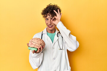 Doctor holding a brain model on yellow studio excited keeping ok gesture on eye.