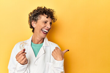 Doctor with invisible dental aligner on yellow points with thumb finger away, laughing and carefree.