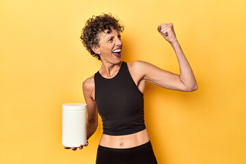 MIddle aged athlete woman holding protein supplement on yellow raising fist after a victory, winner concept.