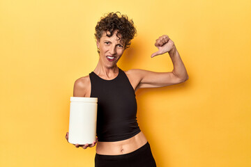 MIddle aged athlete woman holding protein supplement on yellow feels proud and self confident,...