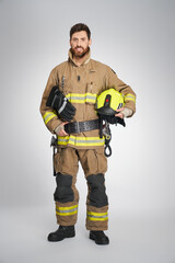Strong, bearded fireman posing to camera with yellow protective helmet. Front view of happy male firefighter keeping hand on leather belt, while looking at camera, on gray background. Concept of work.