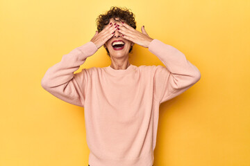 Mid-aged caucasian woman on vibrant yellow covers eyes with hands, smiles broadly waiting for a surprise.