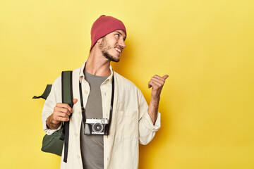 Man with vintage camera and backpack on yellow points with thumb finger away, laughing and carefree.