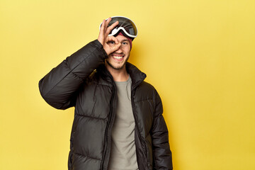 Man in winter coat with snow goggles on yellow excited keeping ok gesture on eye.