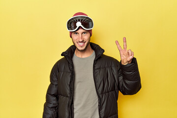 Man in winter coat with snow goggles on yellow showing number two with fingers.