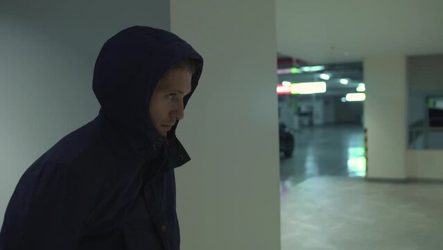 Close-up of a stern-looking suspicious hooded young man look around, hiding from people criminal is guarding the victim in the underground parking