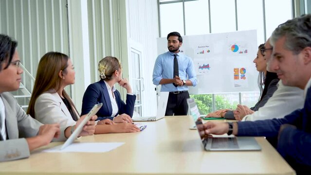 Young indian business manager standing and presenting to group of diversity colleagues in front of white board in meeting at boardroom in office . leader listen and answer questions coworker asking