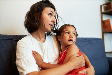 Side view portrait of young mother supporting her child, hugging frightened little daughter on sofa...