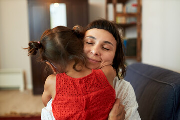 Portrait of mother hugging her daughter in red dress tightly before going to work in morning, standing against door. Mom and little girl embracing after returning home from kindergarten