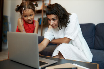 Selective focus on little girl watching her mother working on laptop online as copywriter or...