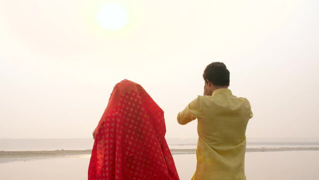Traditionally dressed woman and a man worshiping Lord Sun while standing in a river - celebrating Chhath pooja . A couple standing in a river praying to God Sun - Bihari festival celebration at the...