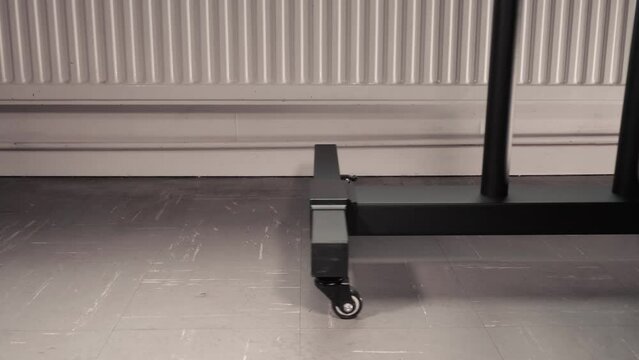 Closeup of the lower part of a generic black metal floor stand on castors, being wheeled into place in front of a heating radiator, in a room with a vinyl tiled floor.