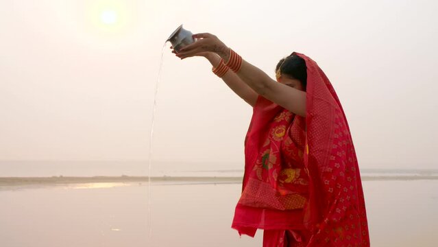A traditionally dressed Indian woman in Chhath pooja attire is worshipping Lord Sun - Hindu festival  offering water. A woman standing in a river giving Argya to Lord Sun - praying to god  celebrat...