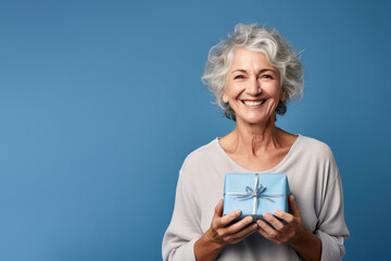 Beautiful mature woman 50, 60, 70 years old on a blue background holding a large gift box, holiday surprise, compliment bonus for birthday anniversary - obrazy, fototapety, plakaty