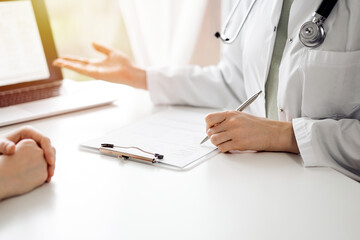 Doctor and patient sitting and discussing something near each other at the white desk in clinic. Female physician is pointing into laptop screen. Medicine concept