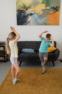 Adorable children looking down while exercising on carpet near sofa set with foot on knee of standing leg and touching raised hands above head in daylight at home