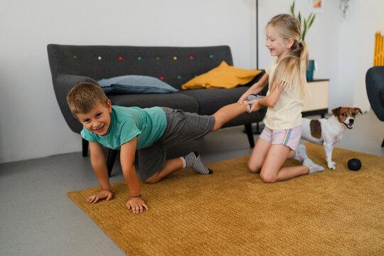 Adorable kid standing on arms knees and looking down while smiling child in ponytail kneeling on carpet near sofa and pulling leg of sibling during play in blurred living room
