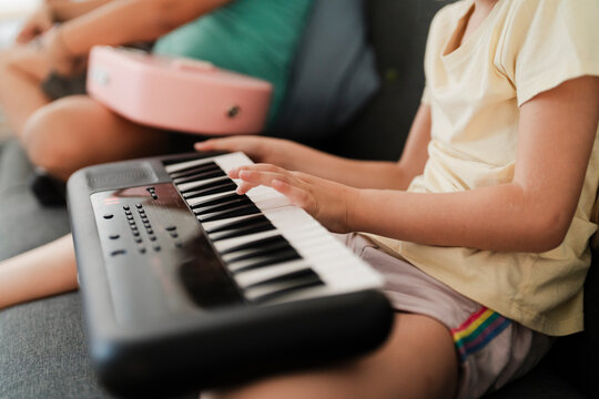Side view of focused girl in casual clothes practicing piano while sitting near blurred boy playing guitar on sofa at home