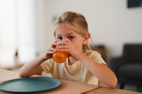 Positive preteen girl in casual clothes with blond hair drinking a glass of orange juice sitting at table looking at camera at home