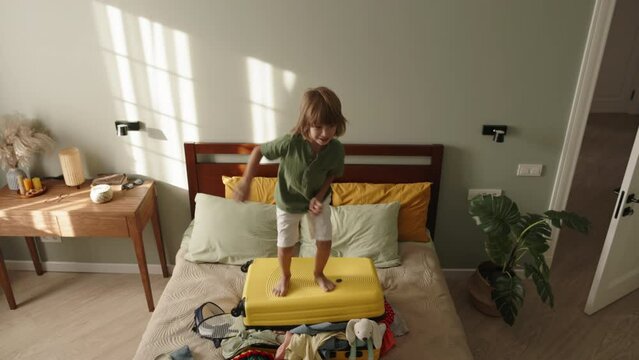 Child's gleeful jumps on packed suitcase, true essence of happy childhood. Unfazed by messiness and randomness of items inside, excitement and anticipation of memorable travel adventure with family