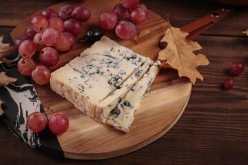 Cheese platter served with autumn fruits and dry leaves on dark wooden background. Appetizer of blue cheese on cutting board with grapes