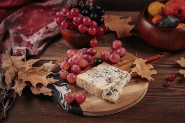 Cheese platter served with autumn fruits and dry leaves on dark wooden background. Appetizer of blue cheese on cutting board with grapes
