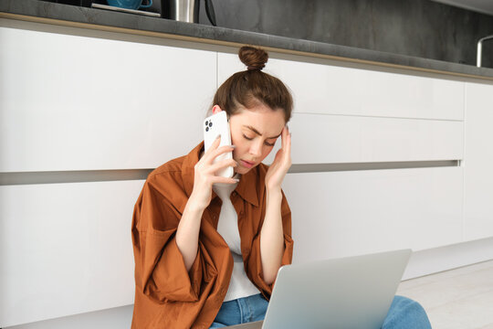 Portrait of worried woman, sits on floor with laptop, listens to bad news over the phone, calling someone with upset, concerned face expression