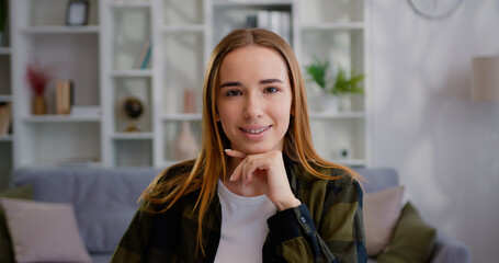 Close up of young beautiful Caucasian woman with long hair beautiful smile looking at the camera while sitting the table against blurred light home or office interior background