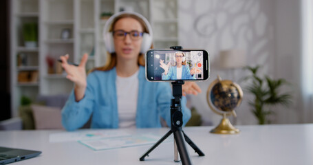 Focus on mobile phone camera from which good-looking confident in headphones young woman recording video vlog for his internet channel from modern light home room
