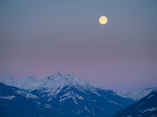 Snowy winter alpine landscape after sunset, pink purple sky with a full moon over a mountain peak