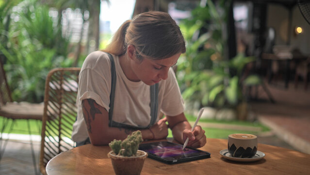 Digital designer draw pic outside cowork cafe. Woman freelancer work outdoor US. Illustrator use tablet stylus pen. Artist paint art sketch pad. Coffee cup. Ipad design app. Talented graphic painter.