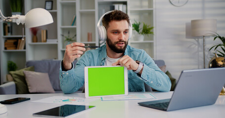 Caucasian man holding green screen tablet while chatting online with partners. Male freelancer using chroma key on her tablet while makes online meeting with colleagues in video call at home