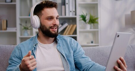 Smiling Caucasian adult man in headphones using digital tablet for e-learning, paying bills online, e-commerce, remote work, social media online at home