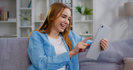 Smiling Caucasian young woman sitting on couch using digital tablet for e-learning, paying bills online, e-commerce, remote work, social media online in living room at home