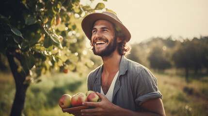 Young adult farmer holding fresh apples, with a beautiful orchard nature landscape in the background, representing a wellbeing, joy, hard work and natural way of living - obrazy, fototapety, plakaty