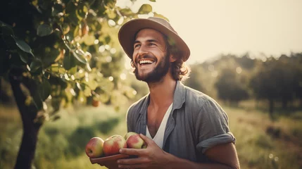 Fotobehang Young adult farmer holding fresh apples, with a beautiful orchard nature landscape in the background, representing a wellbeing, joy, hard work and natural way of living © OTIS STUDIO
