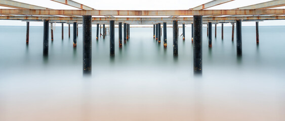 Fine art view of columns under metal pier at sunrise. Shot with long exposure to make the sea appear as fog