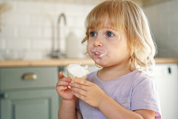 Baby girl enjoying ice cream. Pretty little toddler eating an ice-cream indoors, at home. Dining room background. Small child eats plombir and cream messy on her mouth. Cute kid with tasty sweet food.