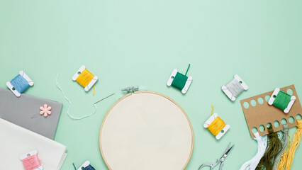 Embroidery set fot stitching. Beige cotton cloth in embroidery hoop on green background with fabric, colorful threads, scissors and needls. Indoor hobby concept with copy space, top view