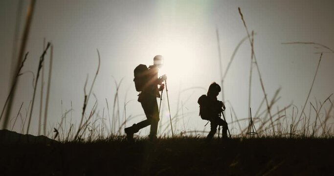 Mother and child conquer mountain, trekking poles backpacks. Bright pleasant sun rays, grass swaying in wind, silhouettes of people, care, succession, exploration, bright moments, love. Family trip