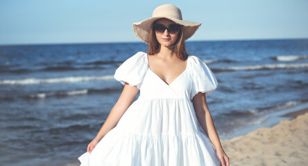 Happy smiling blonde woman is posing on the ocean beach with sunglasses and a hat